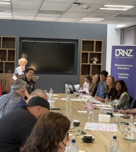 In person Workshop with about 20 staff of Toitu Te Waiora. Pam standing at the front of a long table that has people sitting around it