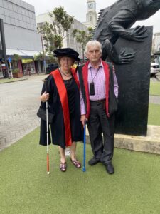 Dr Pam MacNeill, in graduation cap and gown, standing with Don MacNeill in front of a statute